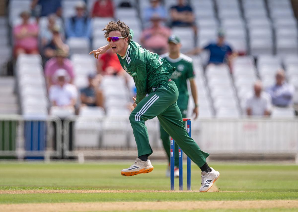 A cricket player in a green uniform bowls during a match, with spectators visible in the background.