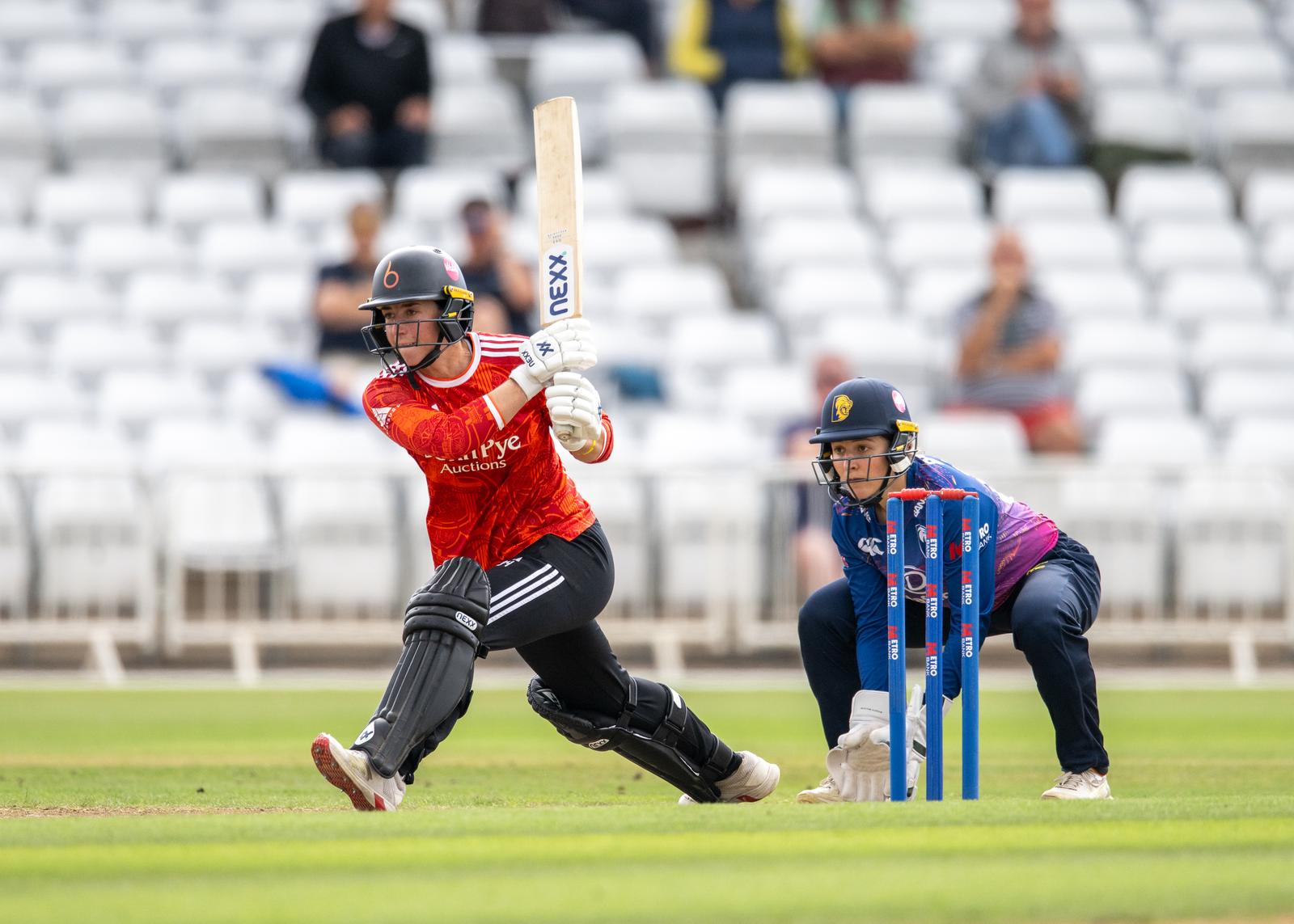 A cricketer in a red jersey swings a bat while a wicketkeeper in blue crouches behind the stumps during a match.